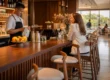 Elegancka lada barowa z drewna dębu z widocznymi słojami, bukowe hokery z tapicerką, barman czyszczący szklanki. Naturalne światło, prestiżowe wnętrze restauracji.